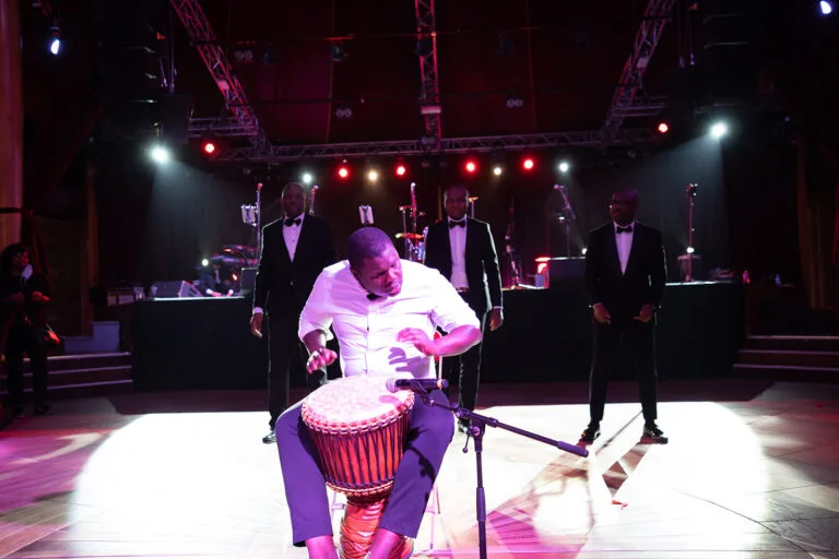 Fran's Orchestra, un percussionniste joue du djembé avec intensité sur une scène éclairée de lumières rouges et blanches, tandis que quatre hommes en costume noir et nœud papillon se tiennent derrière lui.