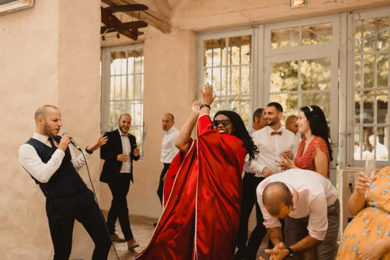 Amany, un moment de fête intense lors d’un mariage. Un chanteur en costume chante avec énergie dans un micro, tandis qu’une femme en robe rouge applaudit en dansant. En arrière-plan, d’autres invités s’amusent et profitent de l’ambiance conviviale sous la lumière naturelle d’une salle aux grandes fenêtres.