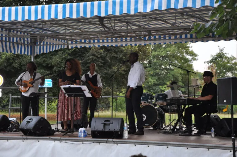 Fran's Orchestra, groupe de musique jouant en plein air sous un chapiteau, avec chanteurs et musiciens interprétant un concert live.