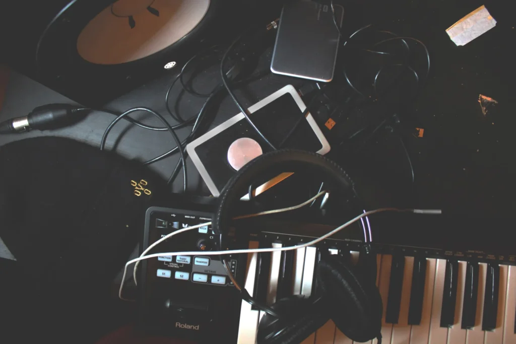 Équipement de studio musical en désordre, comprenant un clavier MIDI Roland, un casque, des câbles et divers appareils électroniques.