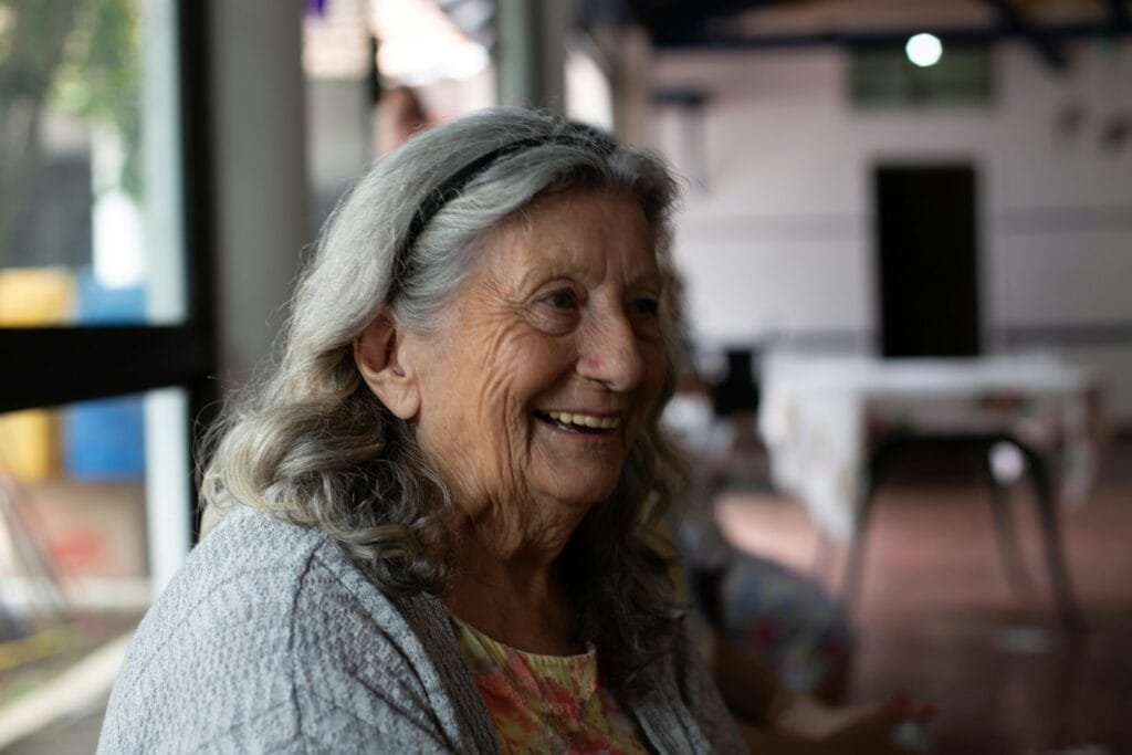 Portrait d’une femme âgée aux cheveux gris ondulés, souriante, portant un bandeau noir et un gilet en tricot, dans un environnement intérieur chaleureux et éclairé.